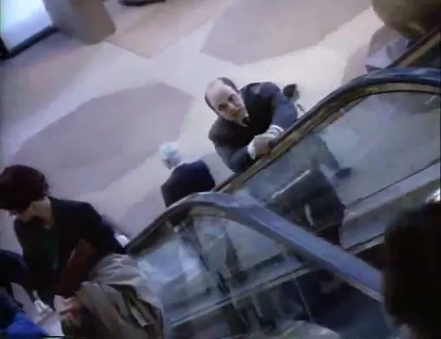 A man in a suit on an escalator looks down at a woman in a trench coat on a lower escalator, as they pass each other in an airport.