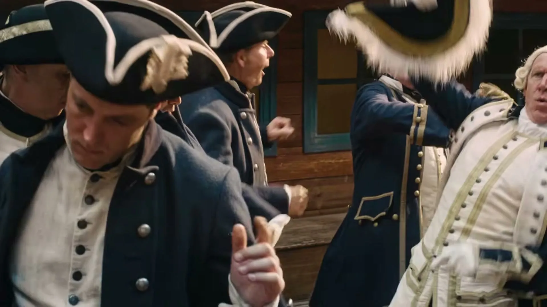 A group of men dressed as 18th-century naval officers are dancing energetically on a wooden deck, with a rustic building in the background.
