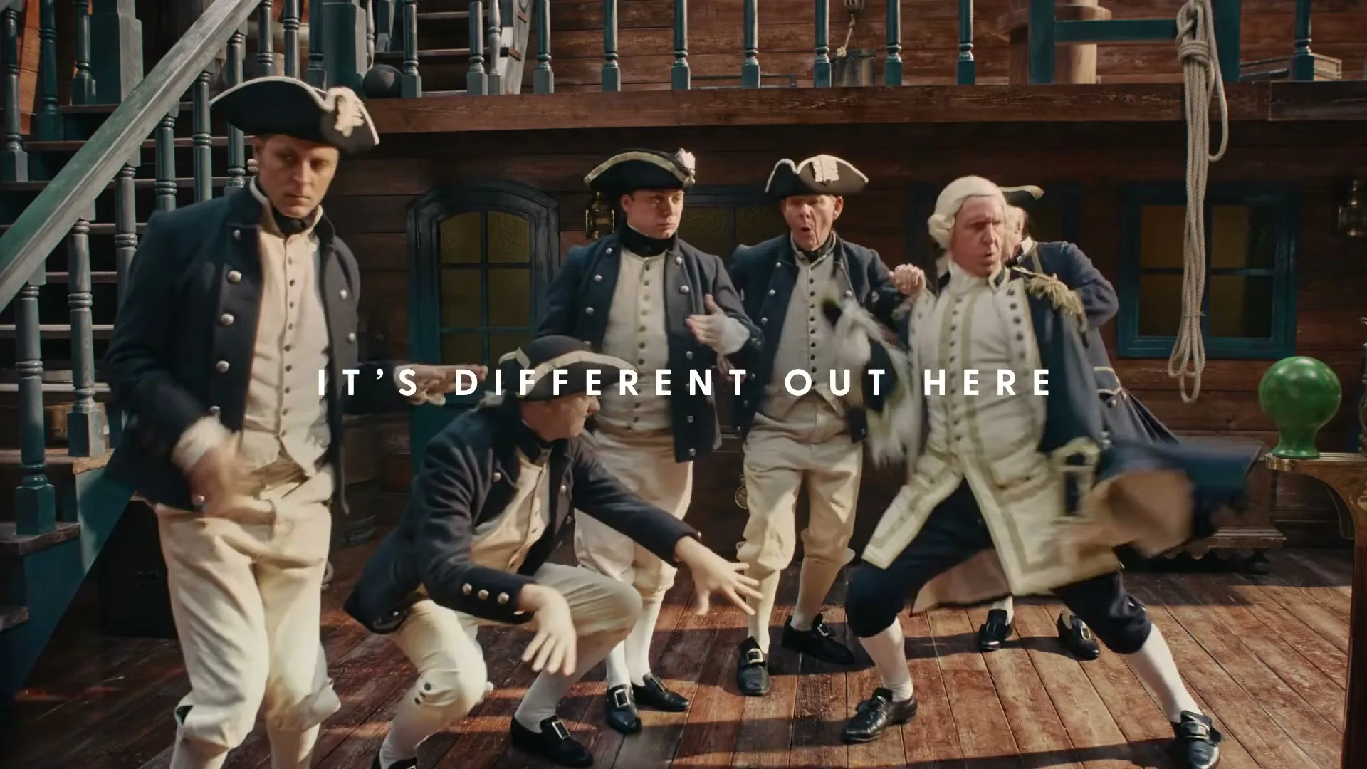 A group of men dressed in 18th-century naval officer uniforms dance energetically on the wooden deck of a ship, with text overlay.