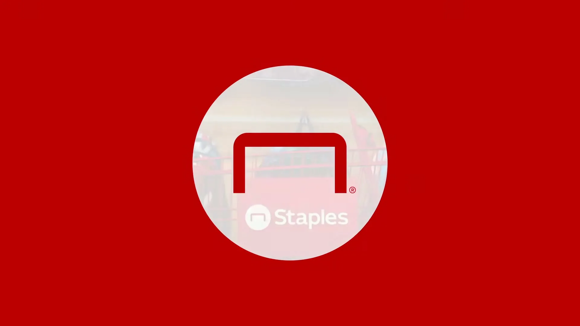 The Staples logo, a red staple on a white circle, is centered on a red background, then fades to reveal a shopping cart and backpacks in a store.
