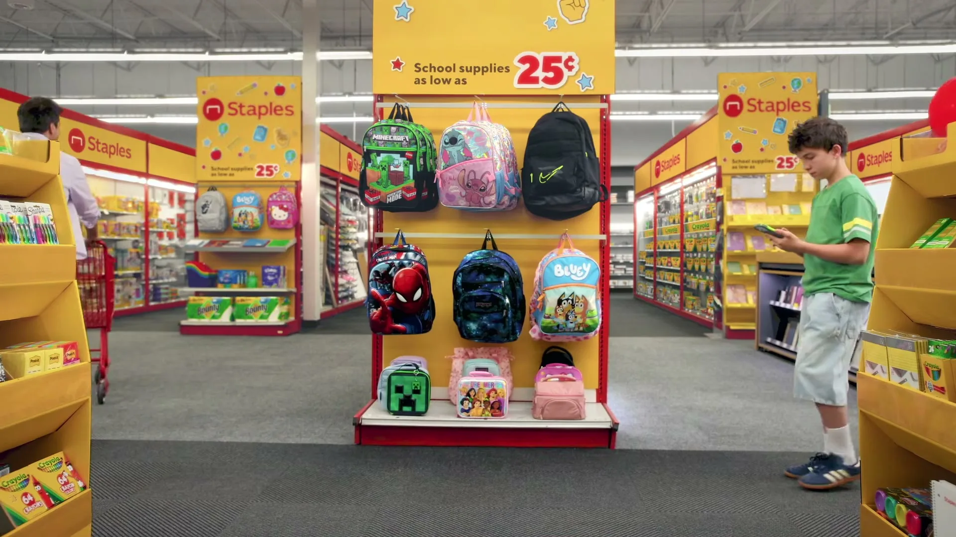 A man pushes a red shopping cart through a Staples store, while a boy looks at his phone and a girl plays with a red balloon.