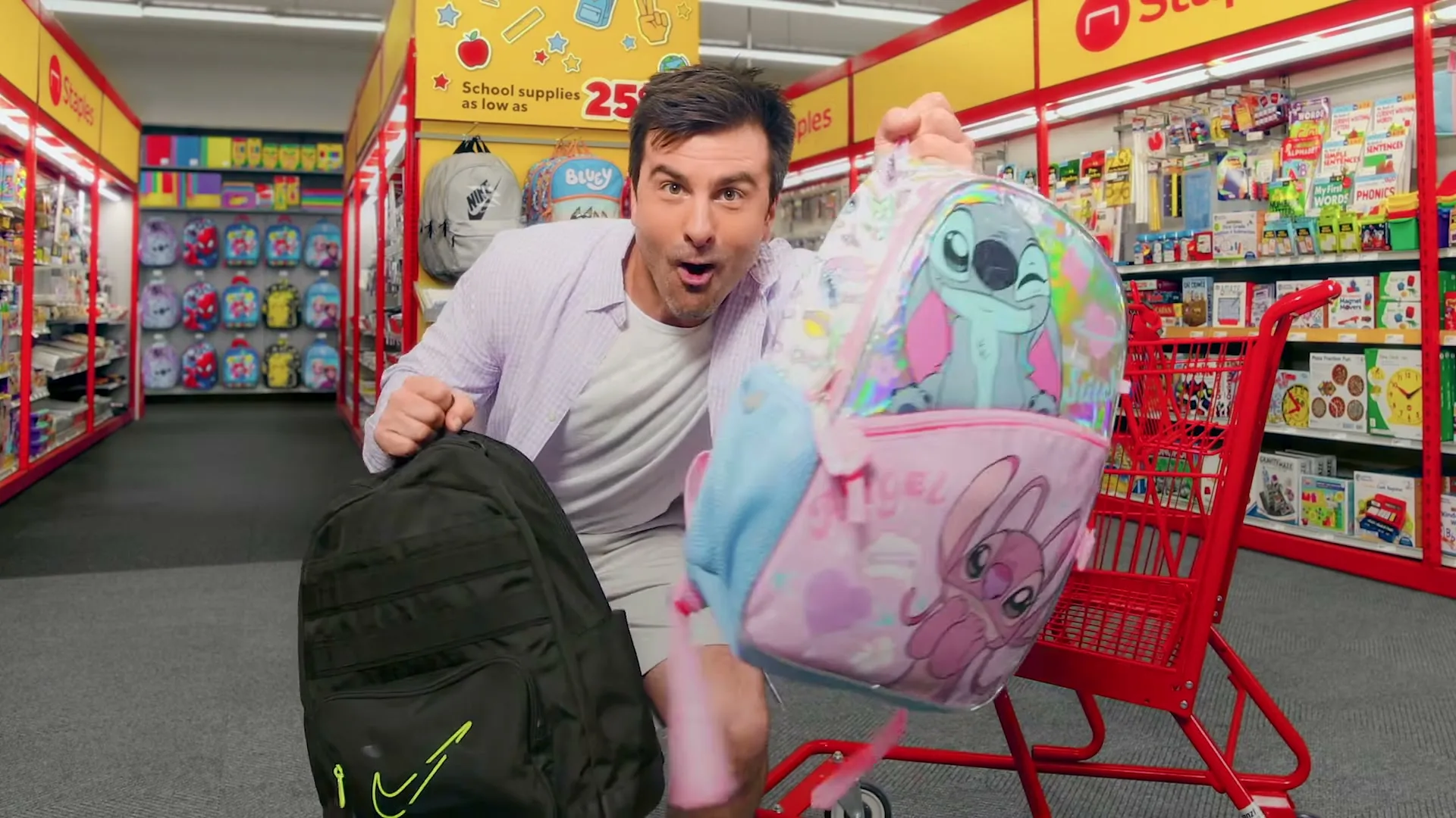 A man in a light shirt and shorts runs excitedly through a Staples store, holding two backpacks and then tossing them into a red shopping cart.