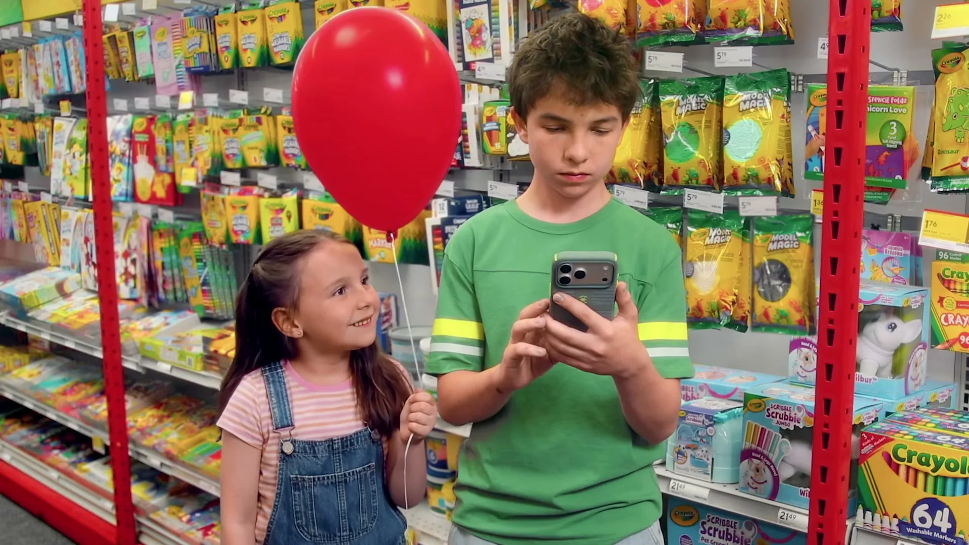 A young boy in a green shirt looks at his phone while a young girl in overalls holds a red balloon and looks at him in a brightly lit store aisle.