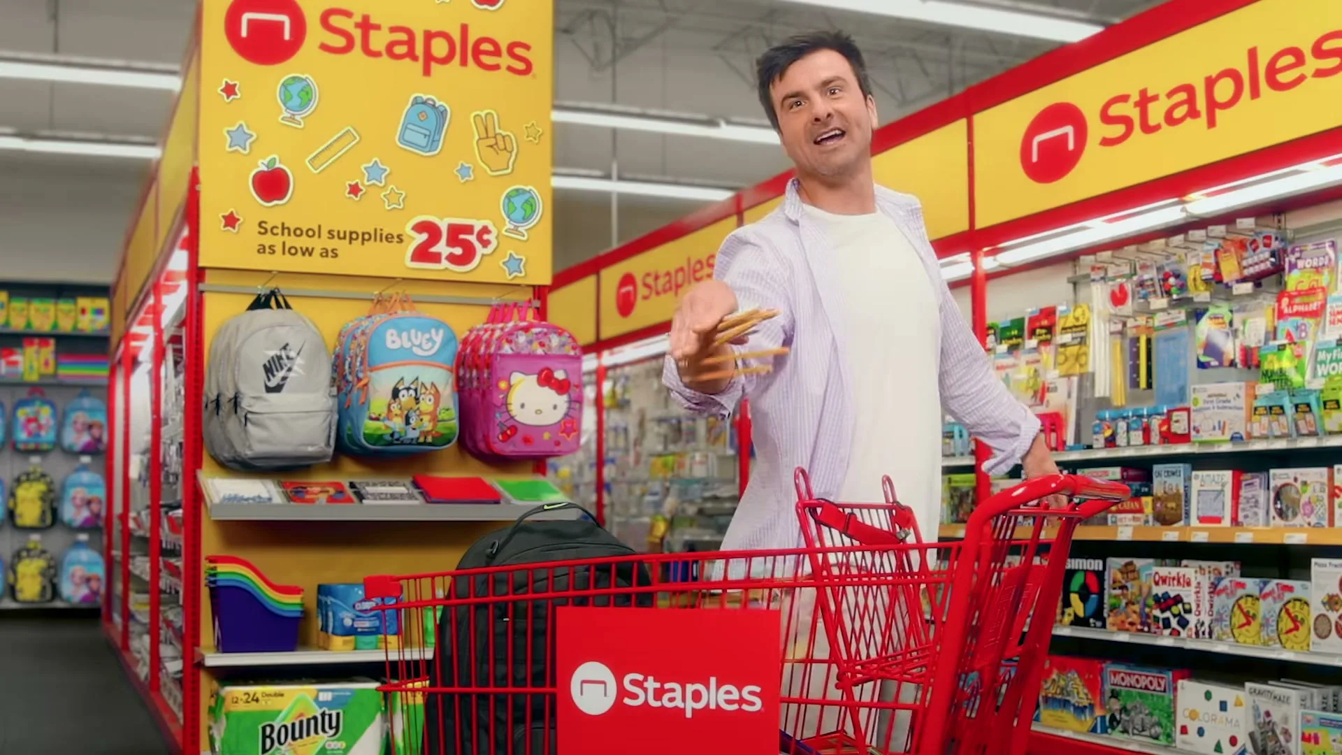 A man in a white t-shirt and purple shirt is pushing a red shopping cart in a Staples store, holding school supplies.