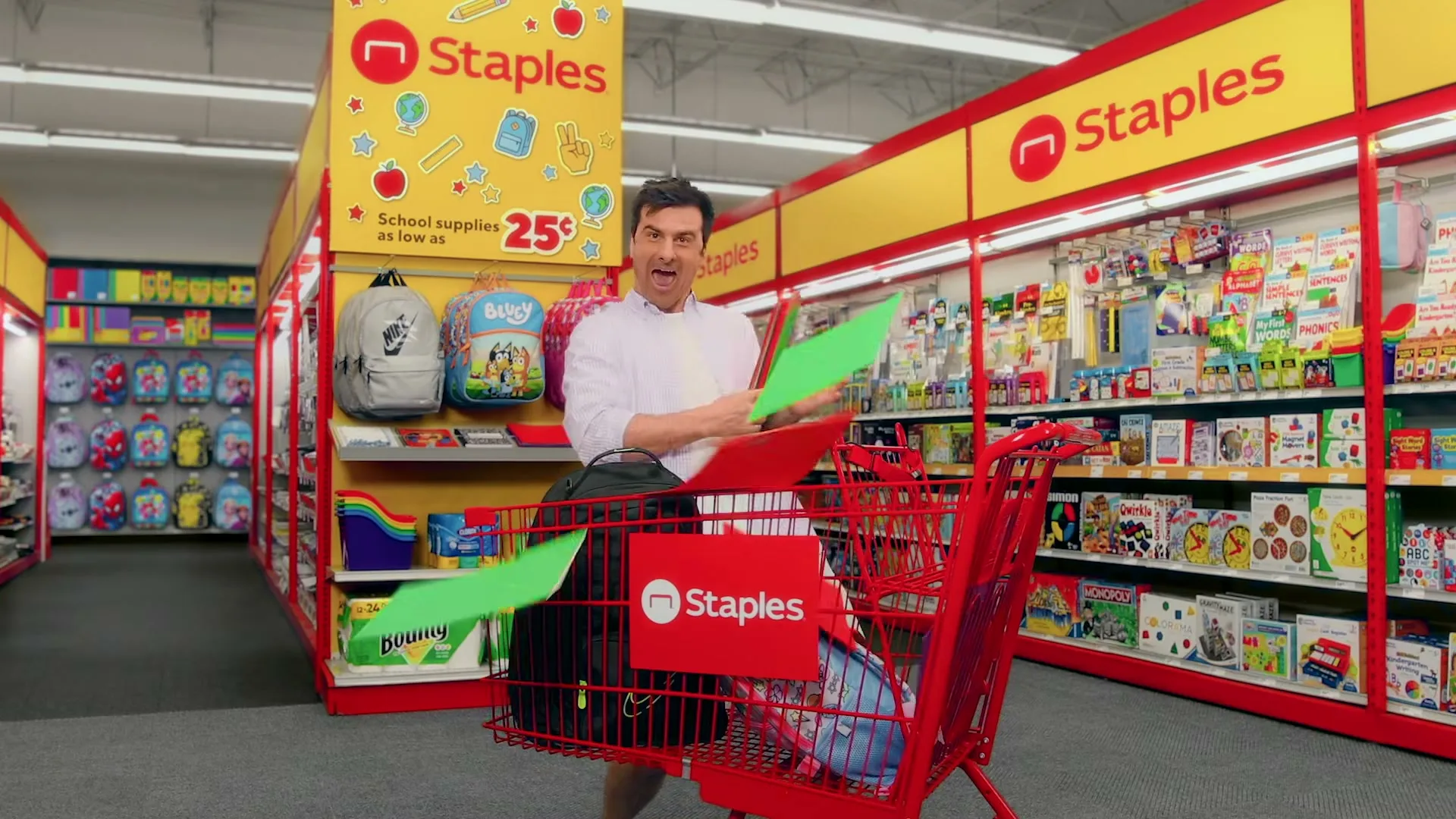 A man in a white shirt dances excitedly in a Staples store aisle, throwing school supplies from his shopping cart.
