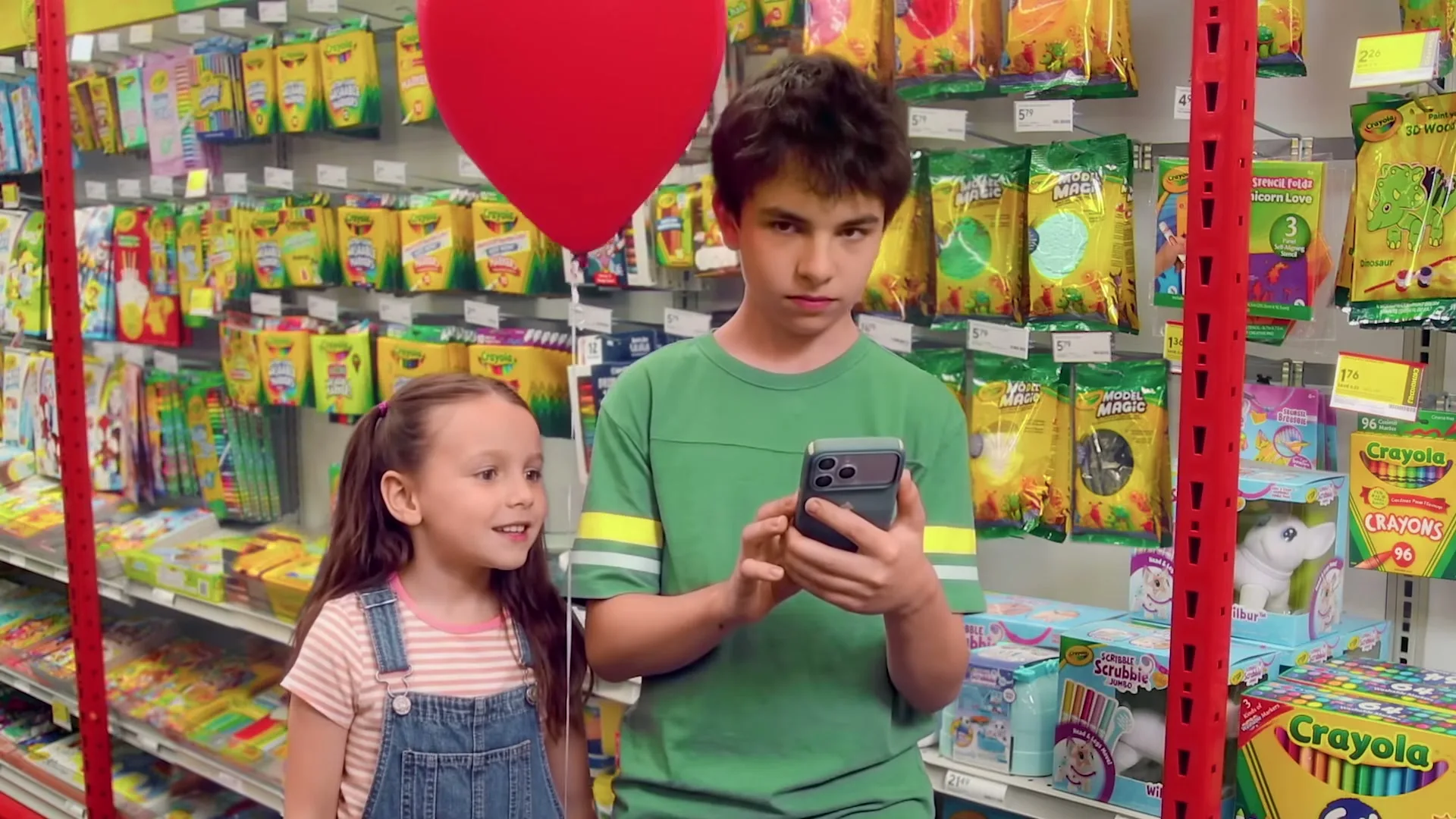 A young boy in a green shirt looks at his phone while a little girl in overalls smiles up at him, holding a red heart balloon in a brightly lit store aisle.