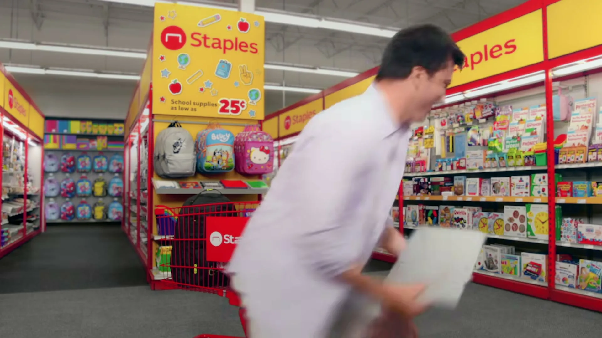 A man in a white shirt and shorts runs through a Staples store, holding a laptop with a "40% off" graphic on its screen, then puts it in a shopping cart.
