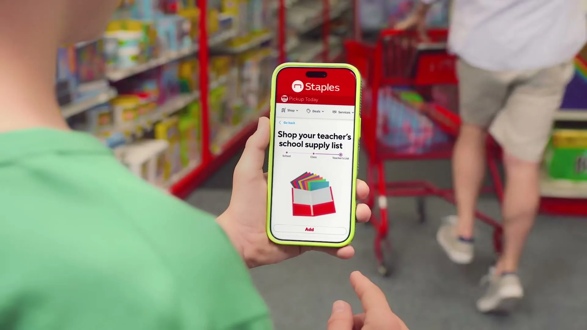 A person in a green shirt holds a smartphone displaying the Staples app, showing a school supply list, inside a Staples store.
