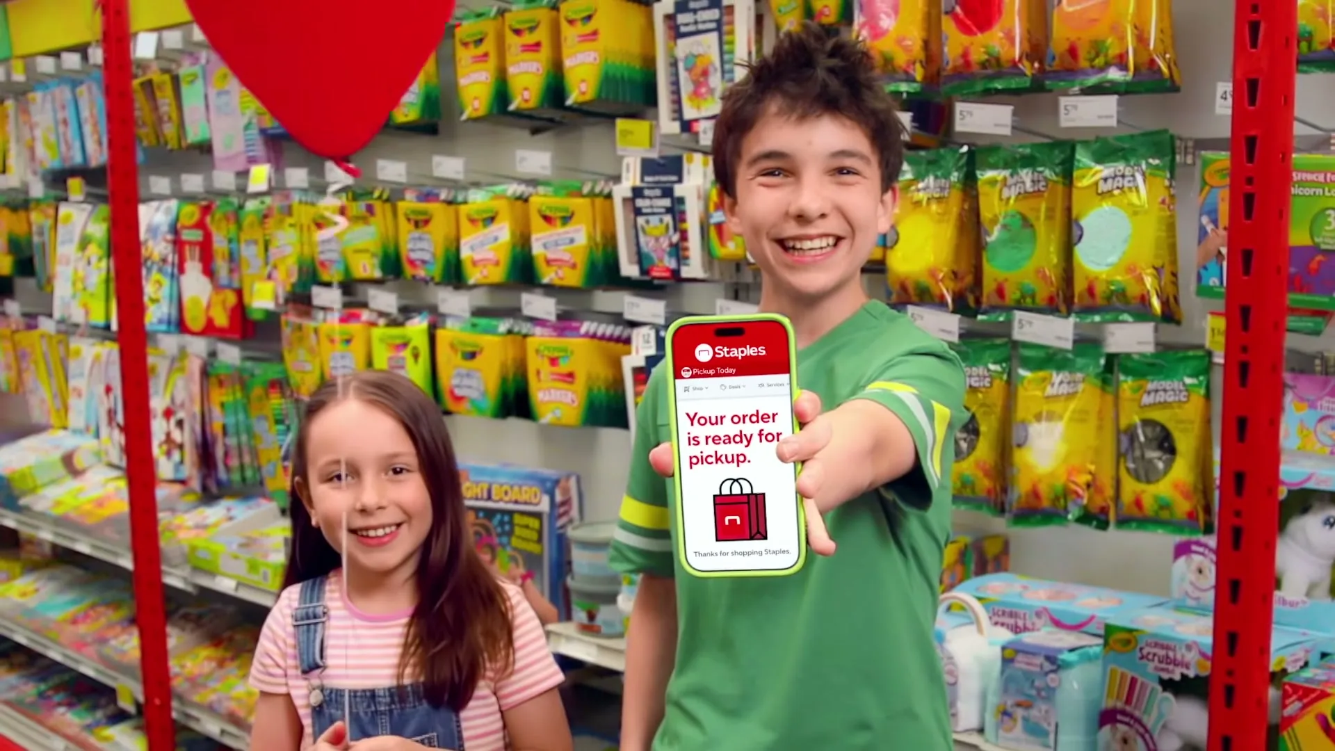 A boy and a girl stand in a Staples store aisle, the boy holding up a phone displaying a Staples app notification for order pickup.