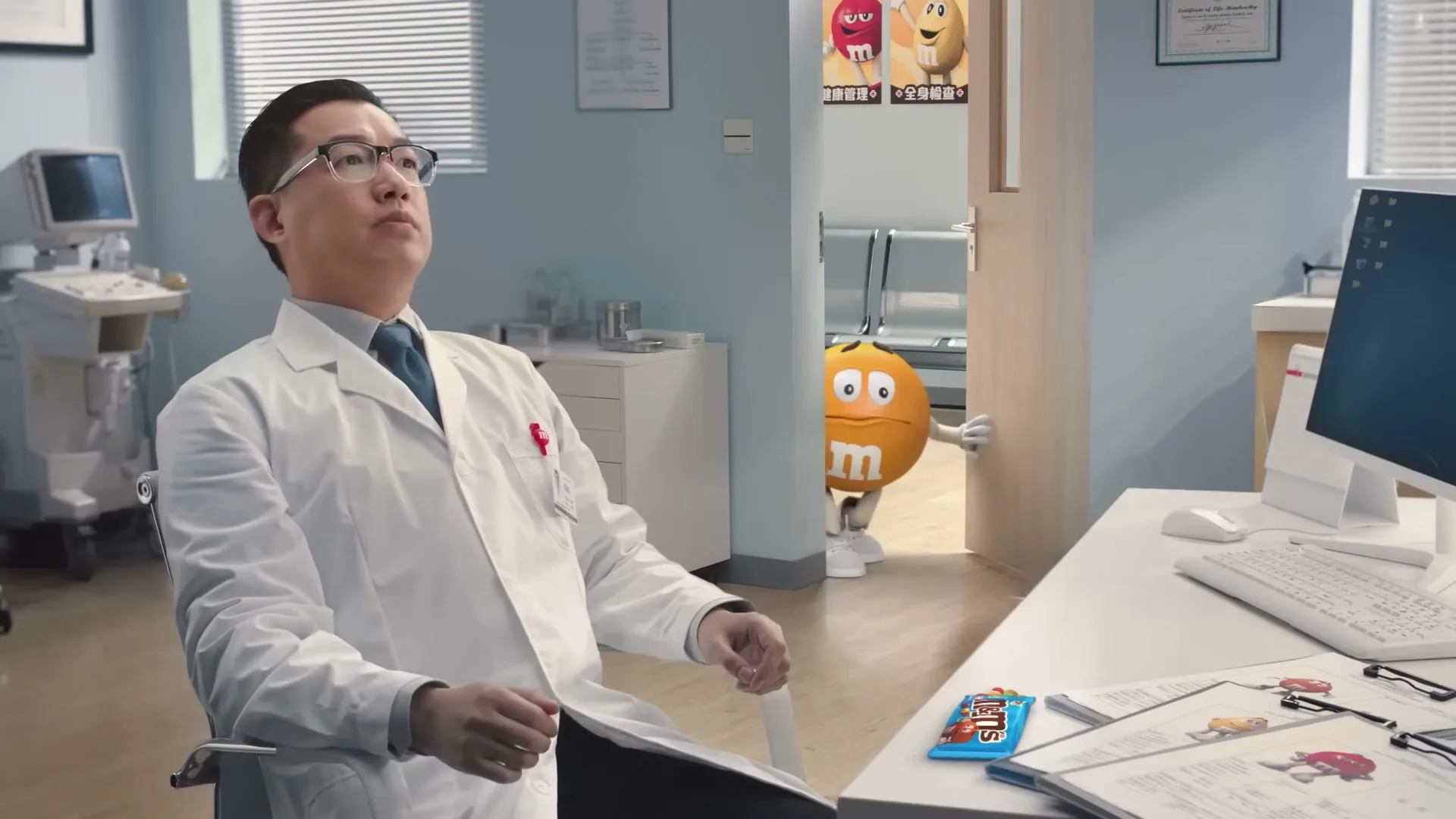 A male doctor in a white lab coat sits at his desk in an examination room, eating an M&M, as the Yellow M&M character peeks from behind a door.