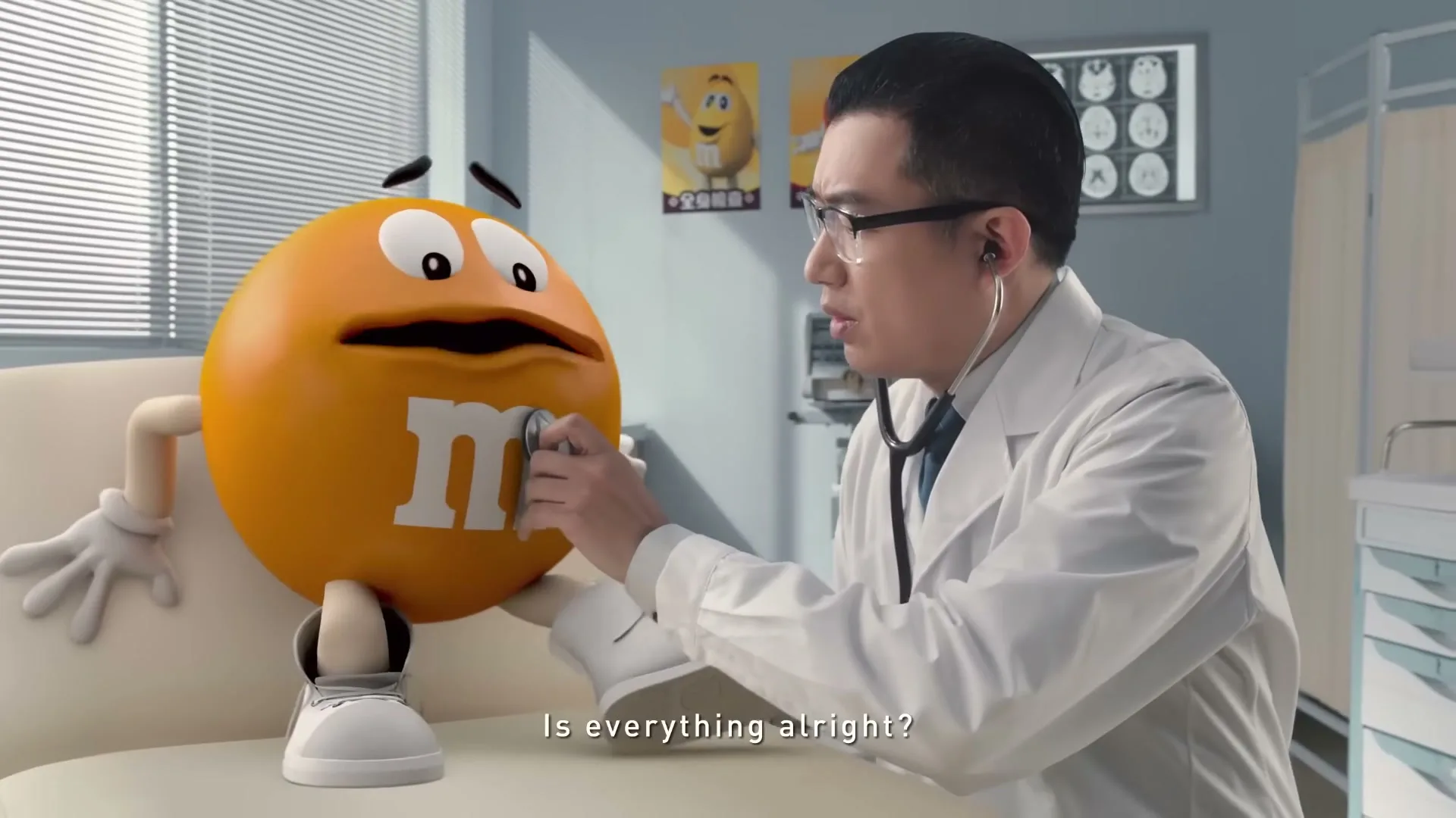 An M&M's character sits on an examination table as a male doctor in glasses listens to its chest with a stethoscope in a doctor's office.