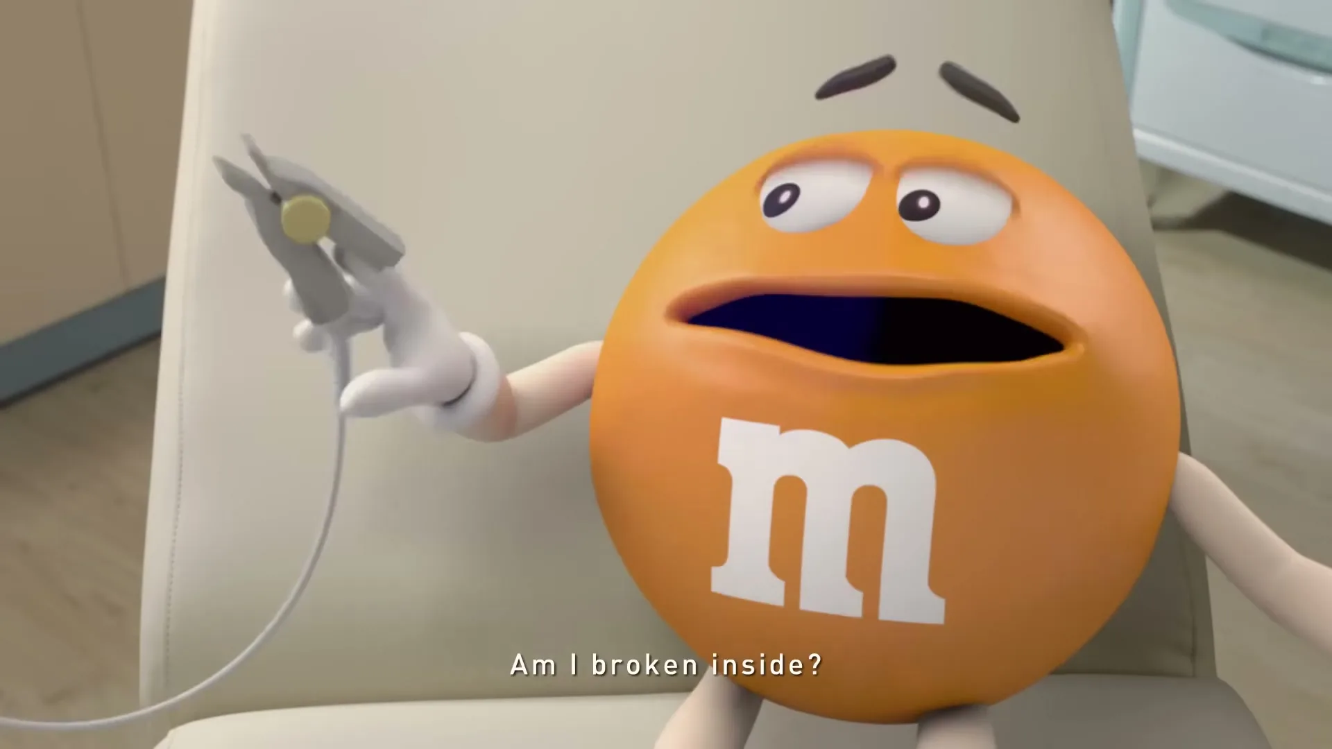 An orange M&M character sits in a chair, looking worried as a human hand holds a medical device to its side in a doctor's office.