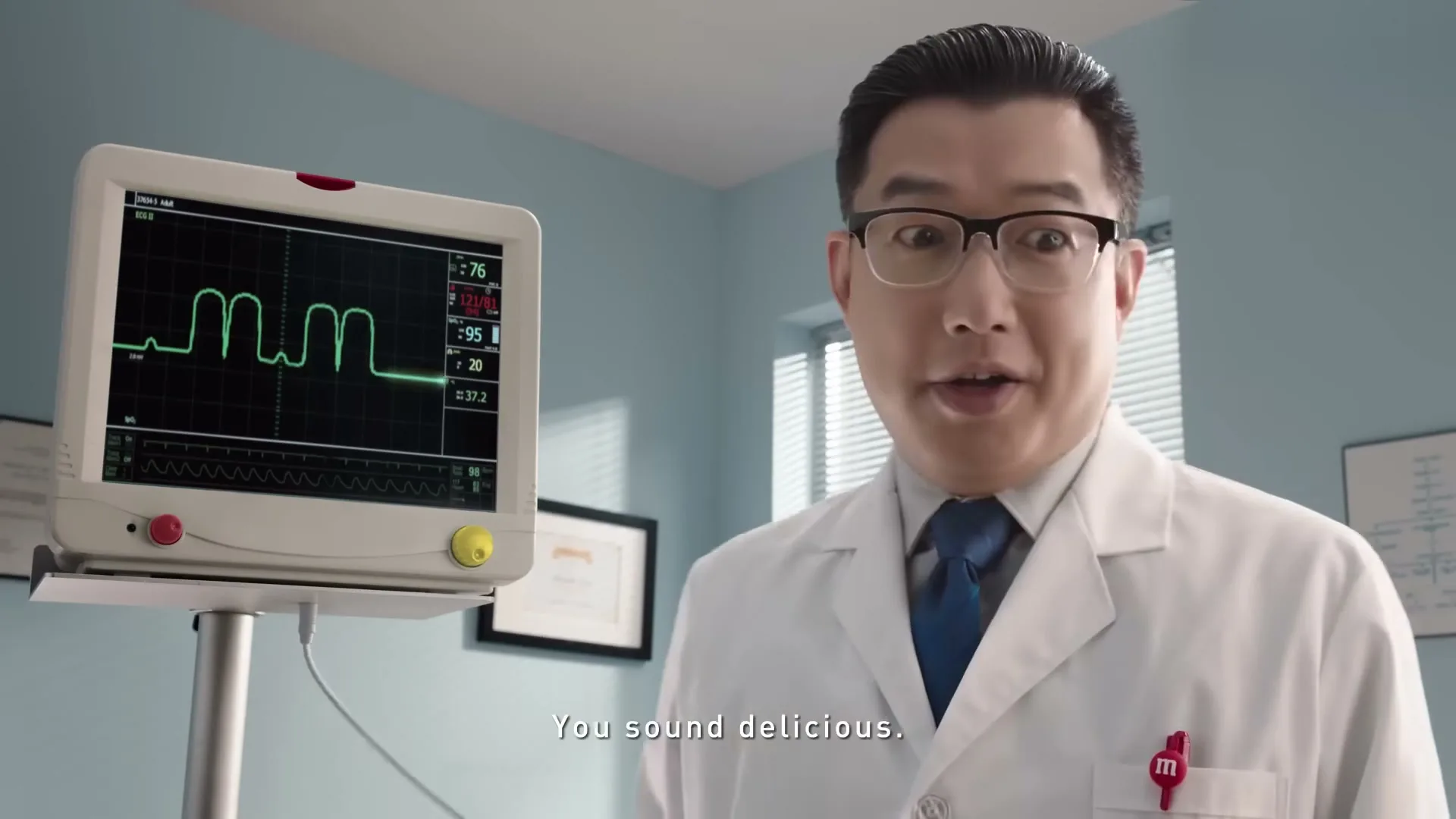 A male doctor in a white lab coat and glasses looks at a medical monitor displaying a wavy line, then turns to the camera and speaks in a doctor's office.