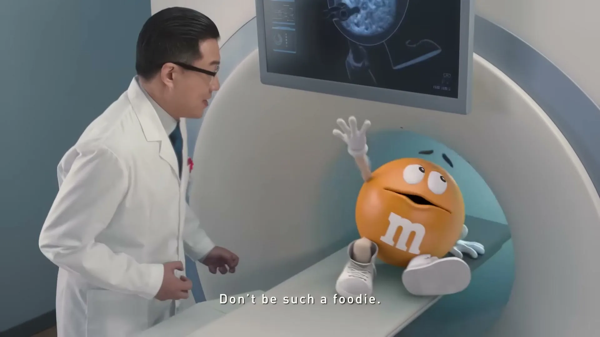 An orange M&M character lies on an MRI machine while a male doctor in a lab coat looks at him and a screen showing the M&M's crispy interior.