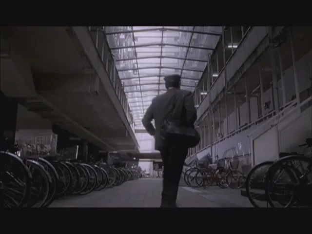 A man in a dark suit and hat runs through a long, covered bicycle parking area with a glass roof, disappearing into the distance.