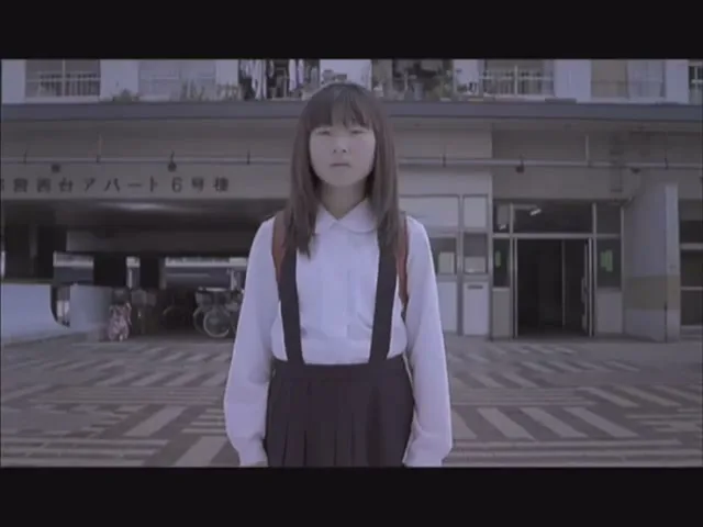 A young Japanese girl with a backpack stands in front of a building, looking directly at the camera with a serious expression.