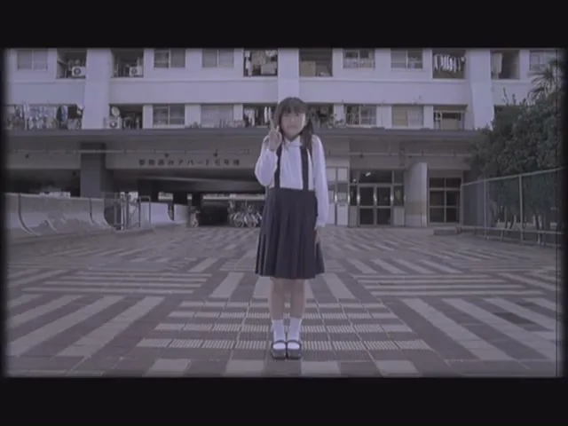 A young Japanese girl in a school uniform stands still in front of an apartment building, holding a phone to her ear.