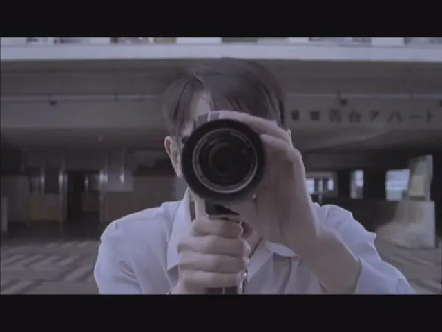 A man with glasses and a white shirt holds a video camera, smiling at the viewer, then lowers the camera to film.