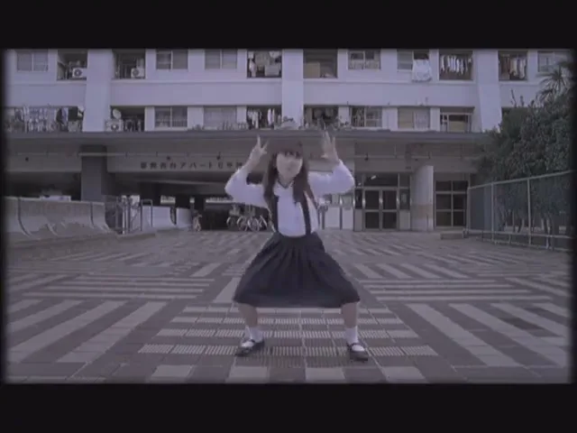 A young Japanese girl in a school uniform dances energetically in front of a large apartment building on a paved plaza.