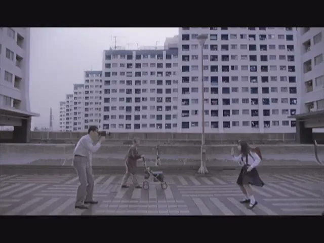 A man photographs a young girl in a school uniform, while an older woman pushes a stroller between them, in an urban setting with large apartment buildings.