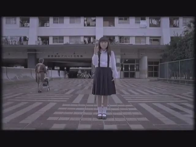 A young Japanese girl in a school uniform stands still in front of an apartment building, holding up a peace sign.