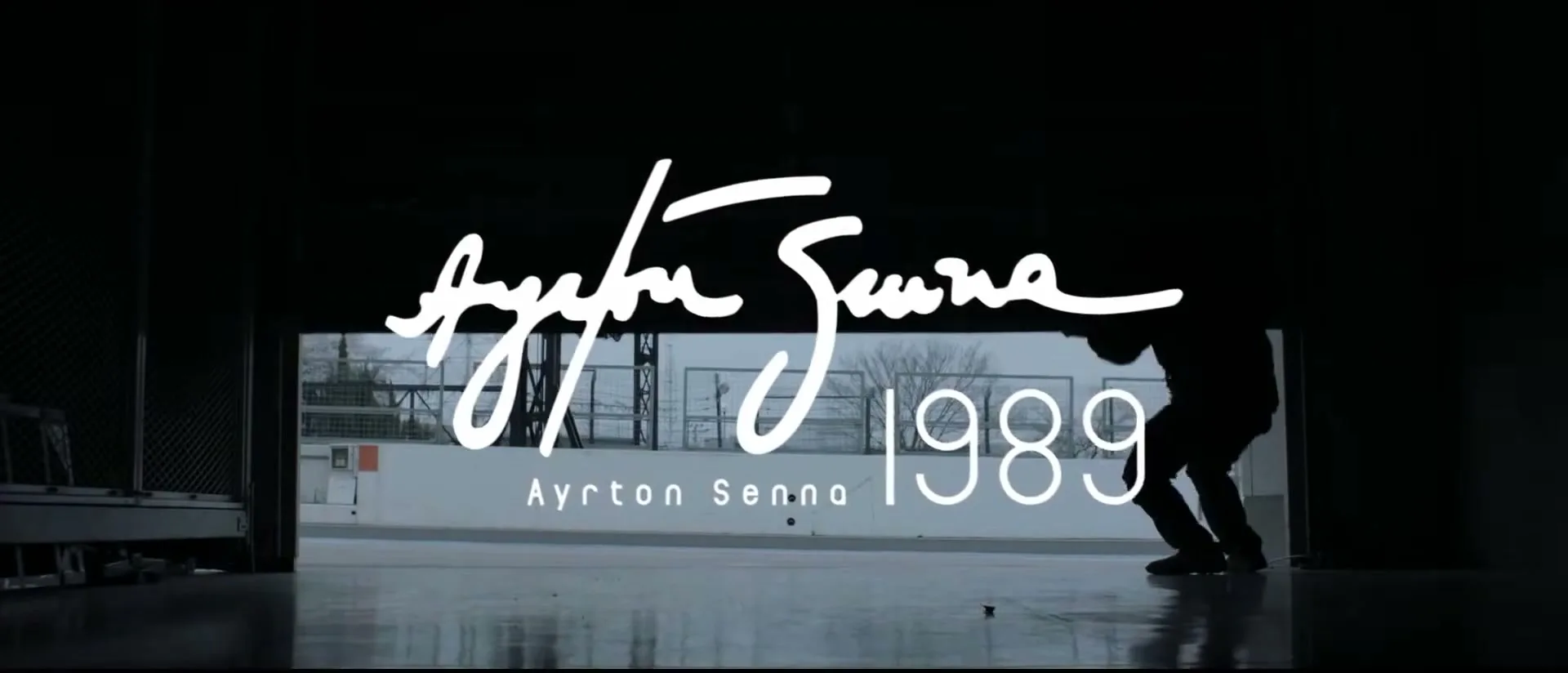 A person in silhouette lifts a garage door, revealing a grey sky and industrial background, with "Ayrton Senna 1989" text overlaid.