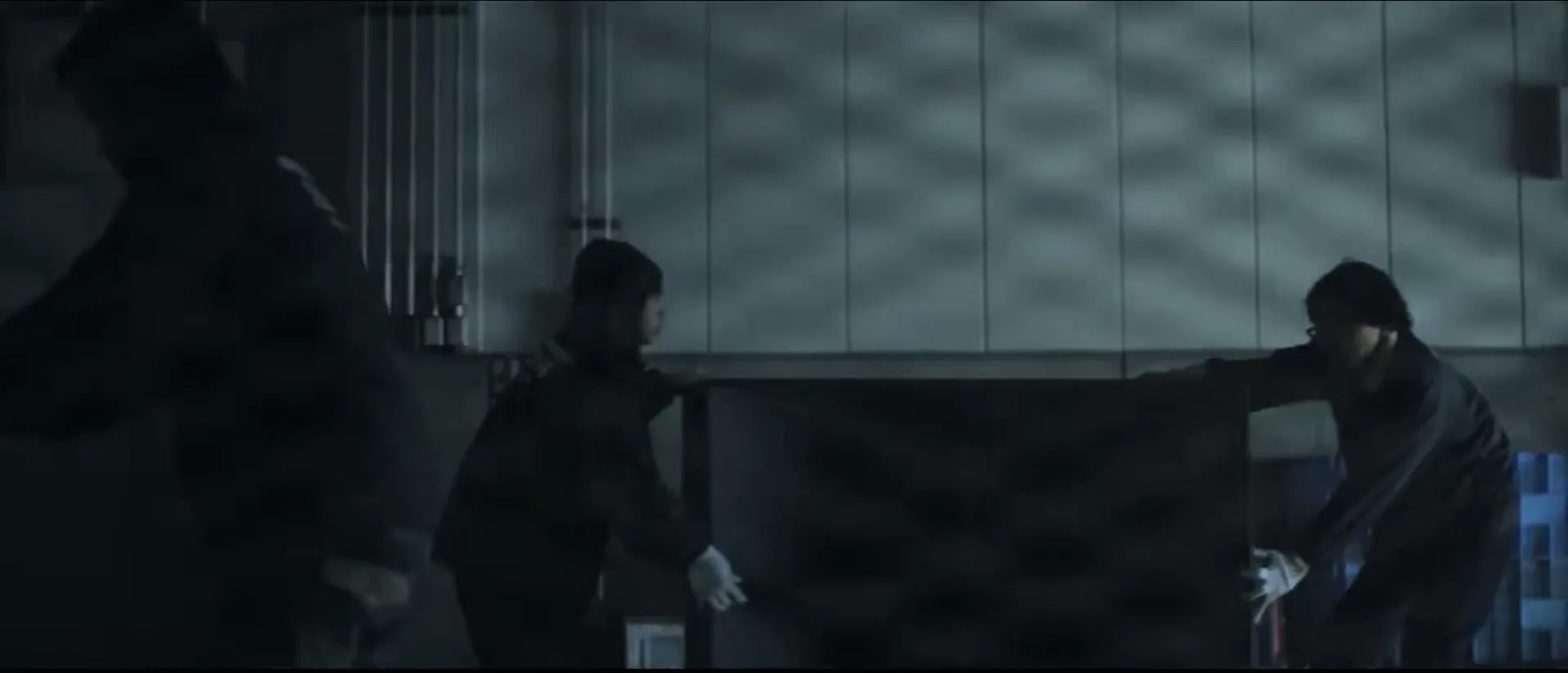 Three men in dark clothing and white gloves carry a large, dark panel through a dimly lit, industrial-looking room.