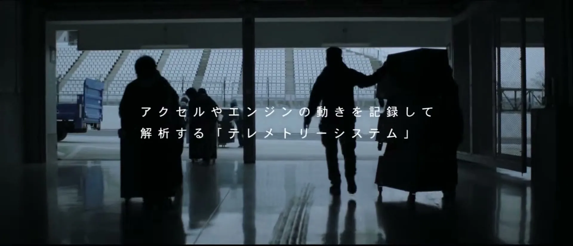Silhouetted figures of a pit crew push equipment through a garage towards a race track, with Japanese text overlaid.