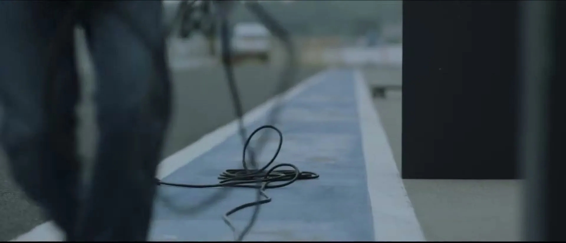 A person in dark pants walks along a blue and white striped track, dragging a black cable behind them, at a race track.