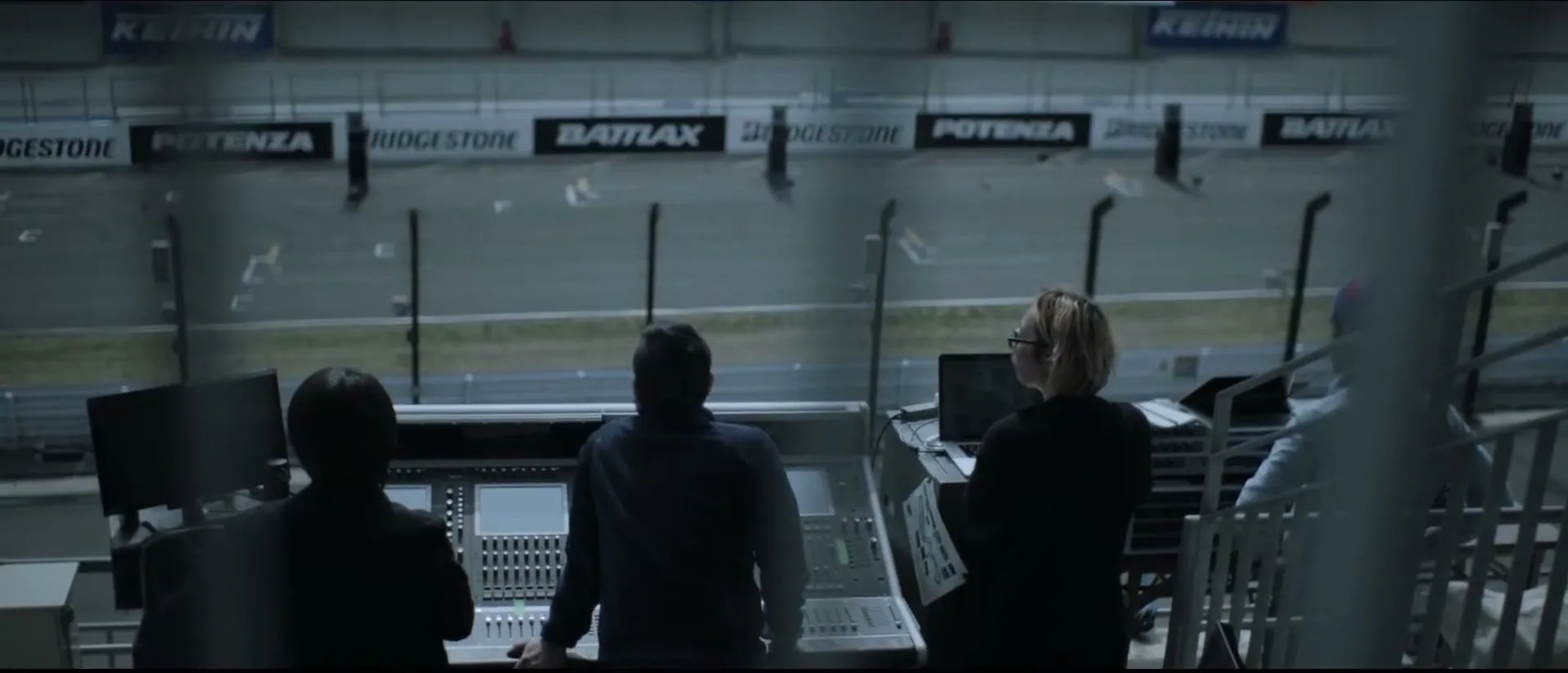Three people, two men and one woman, stand with their backs to the camera, looking out over a race track from a control booth.