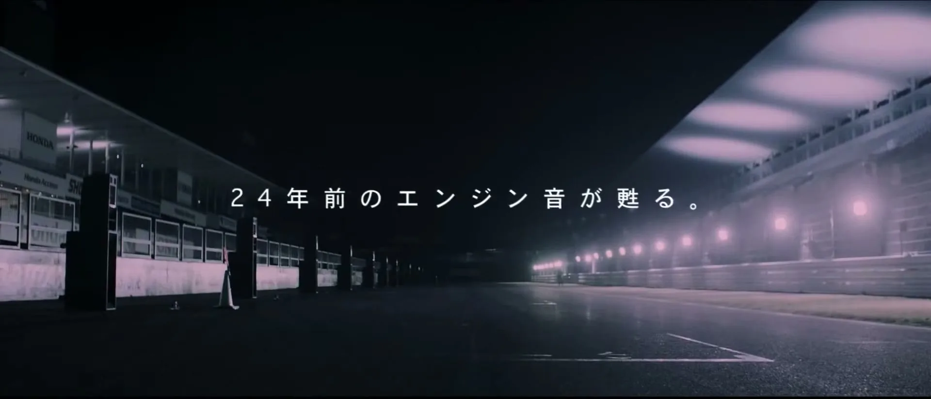 A wide shot of an empty, dark race track at night, illuminated by purple-hued lights, with Japanese text appearing on screen.