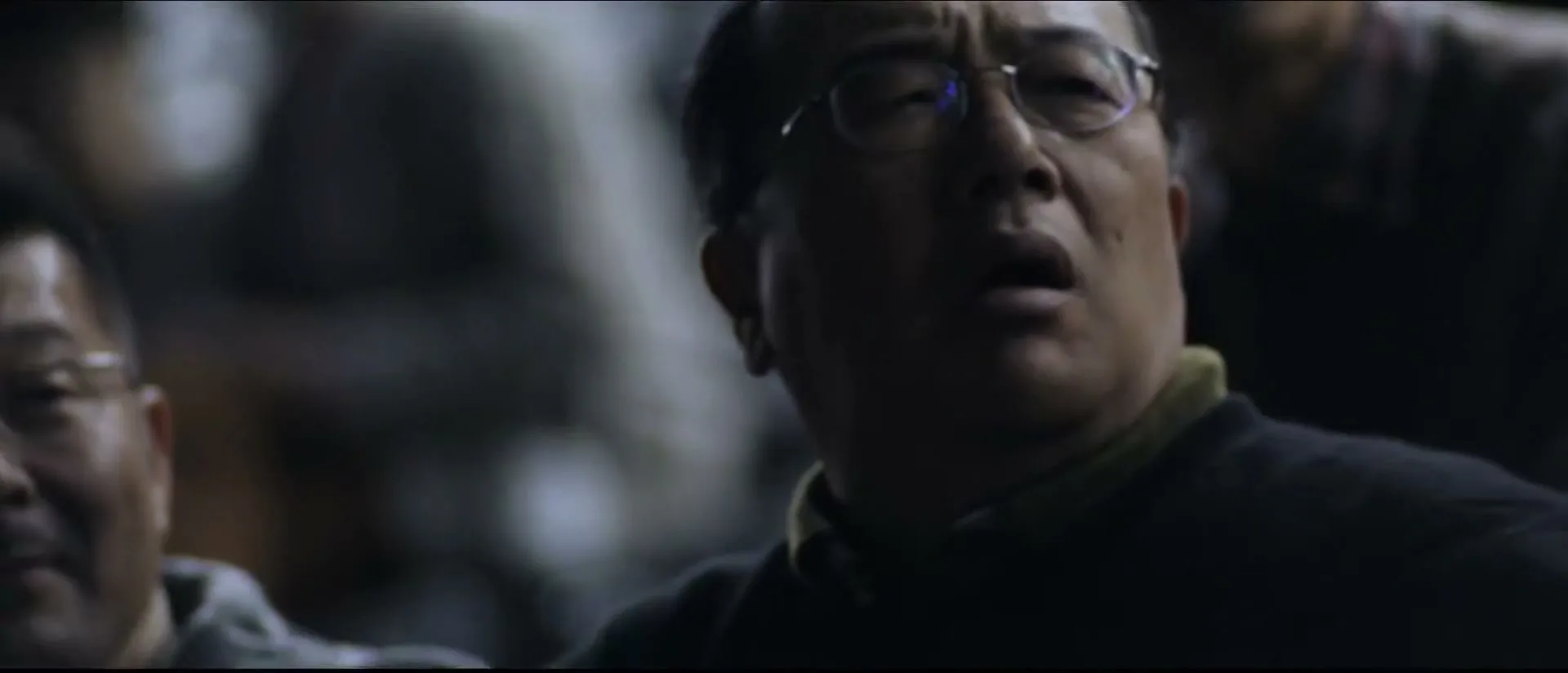 A middle-aged East Asian man with glasses looks up in awe, surrounded by other people in a dark, indeterminate interior space.