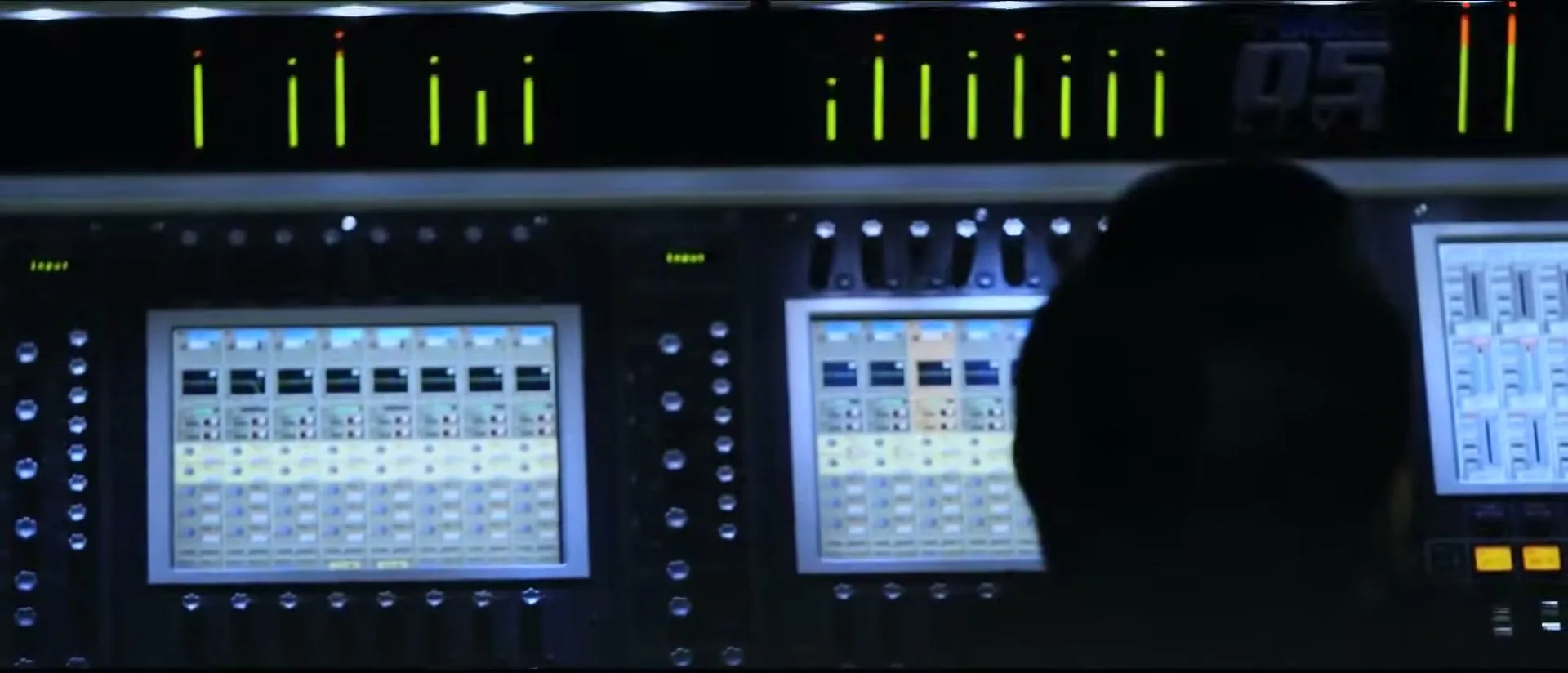 A person's silhouette is seen from behind, looking at a large sound mixing board with glowing green lights and multiple screens in a dark room.