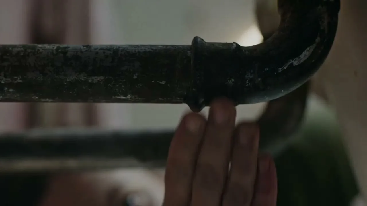 A close-up shot shows a rusty pipe with a single drop of water leaking from it, as a person's hand reaches up to touch the leak.