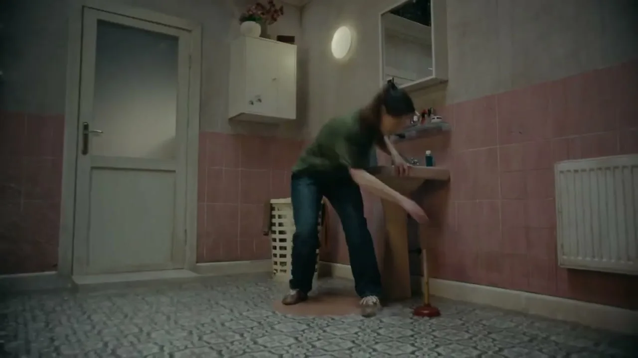 A young woman in a green shirt and jeans struggles to unclog a sink with a plunger in a pink-tiled bathroom.