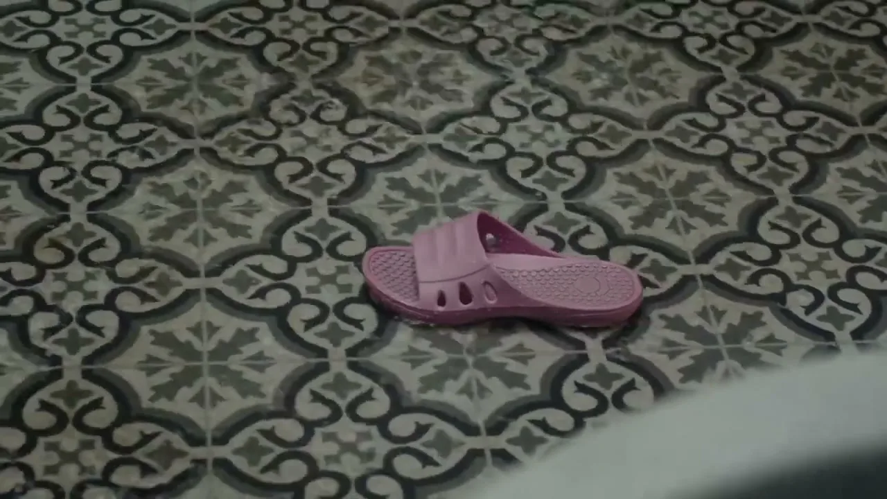 A single pink shower sandal lies on a patterned tile floor in a bathroom.