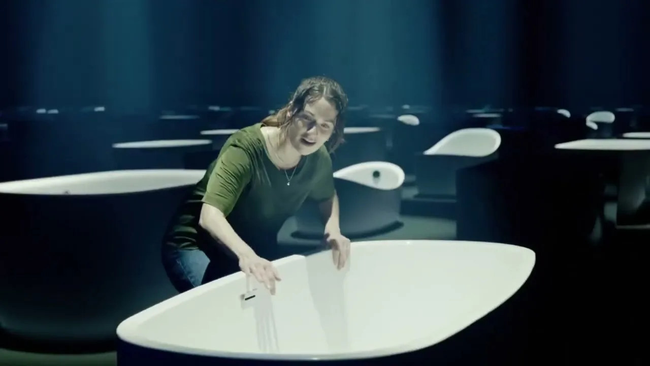 A woman in her 30s, wearing a green shirt and jeans, smiles while holding a white bathtub in a dark, vast showroom filled with many bathtubs.