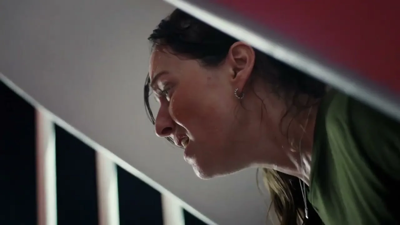 A woman with dark hair and a green shirt, sweating and looking distressed, is seen from a low angle, partially obscured by a white and red structure.