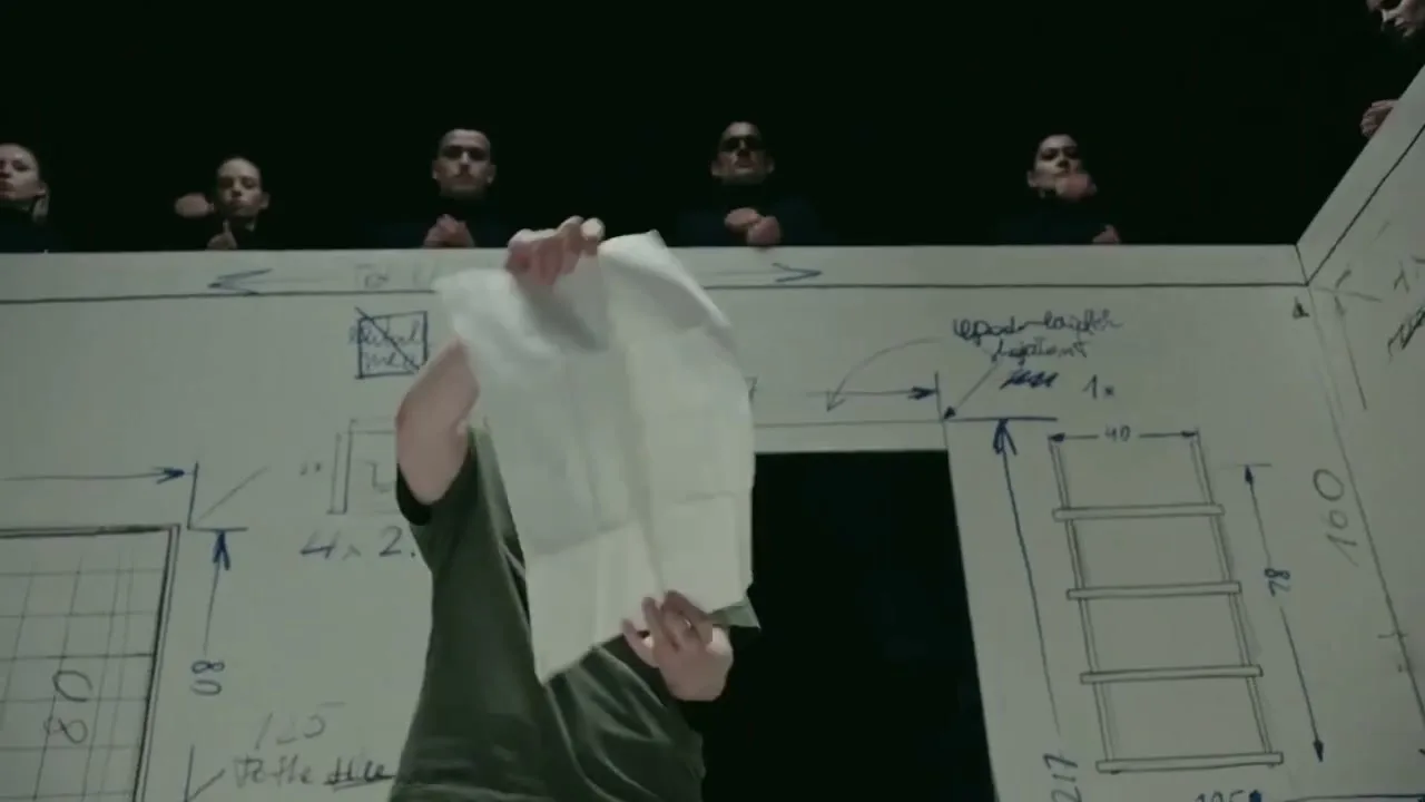 A woman in a green shirt holds up a piece of paper, obscuring her face, while a group of people watch from above in a room with architectural drawings on the walls.