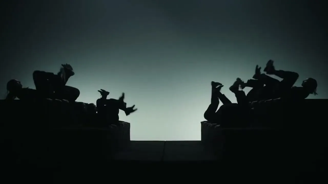 Silhouetted people on two ledges, facing each other, kick their legs in a synchronized, dramatic fashion against a stark, light background.