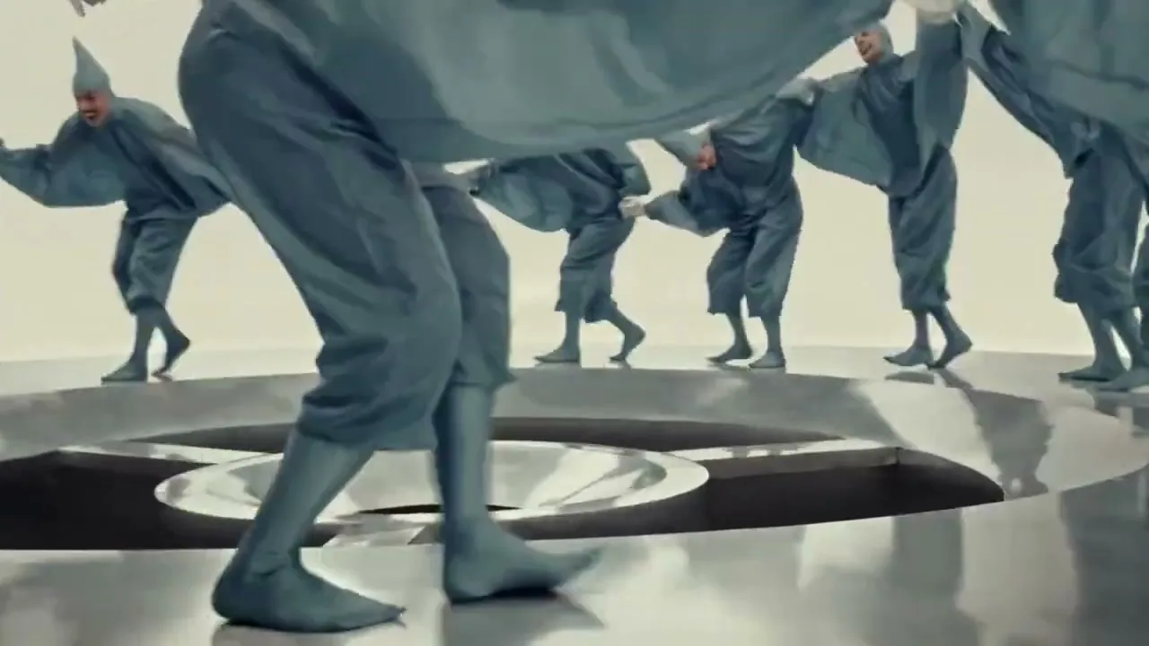A group of people in grey, flowing costumes and pointy hats dance in a circle around a metallic, funnel-like structure in a bright, minimalist setting.