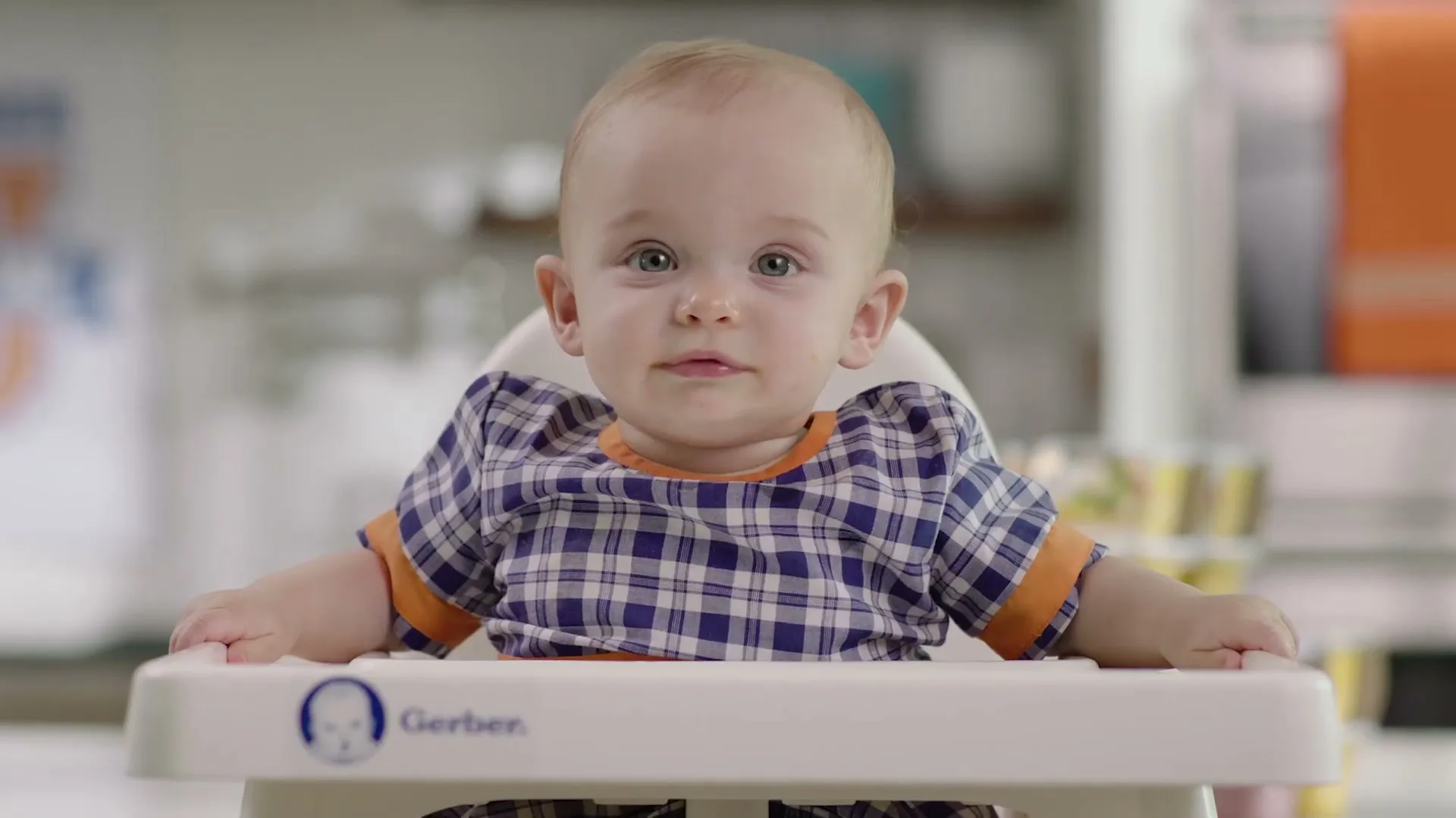 A happy baby boy with light hair and blue eyes sits in a high chair, looking around and babbling in a bright kitchen.