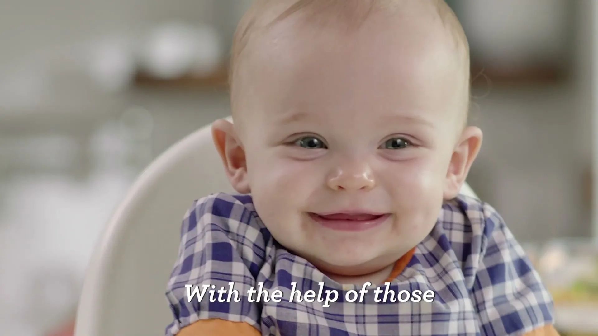 A cute baby with blue eyes and a plaid shirt sits in a high chair, looking around and smiling in a bright, natural kitchen setting.