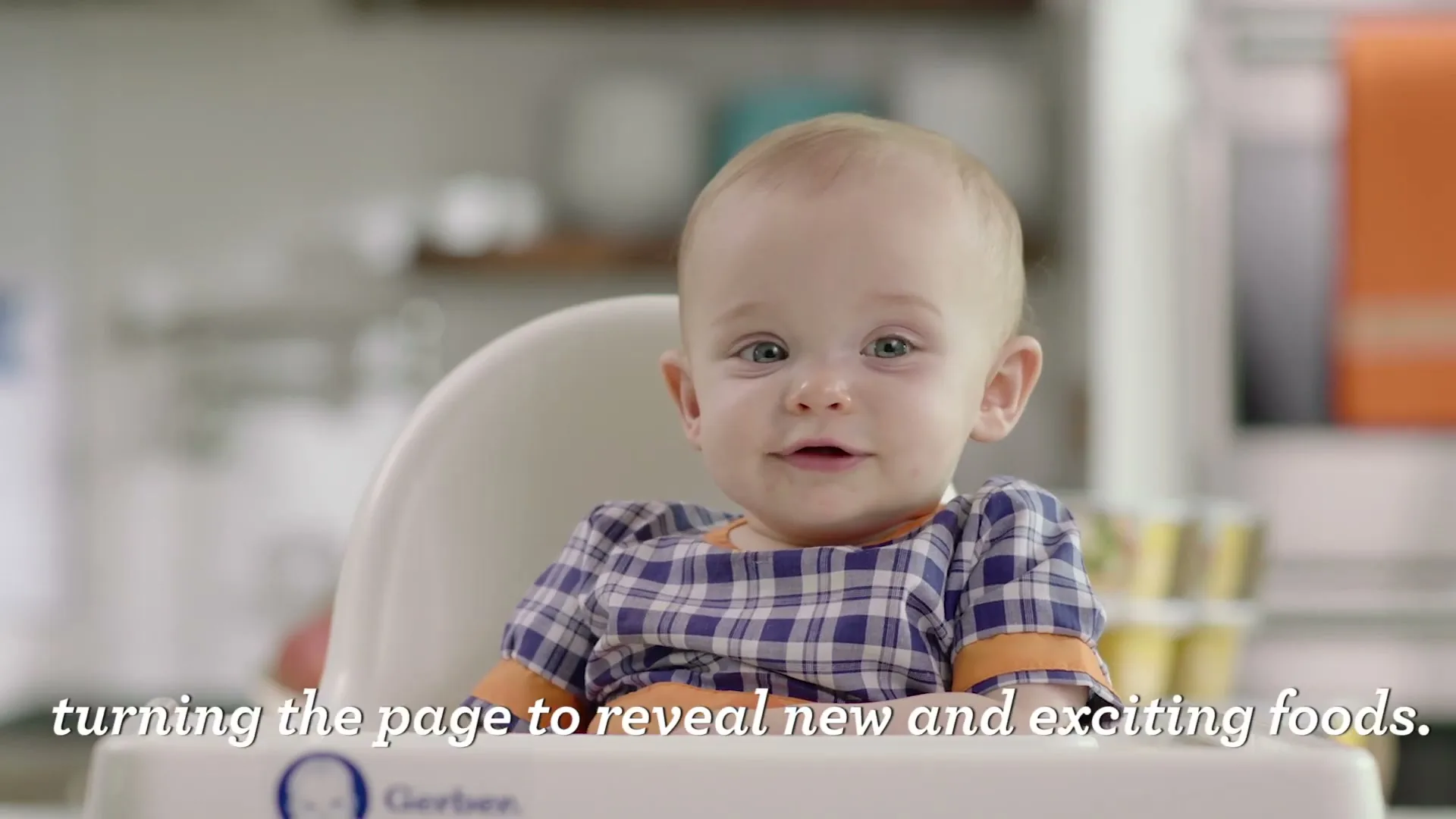 A cute baby in a plaid shirt sits in a high chair, looking around and smiling in a bright kitchen.
