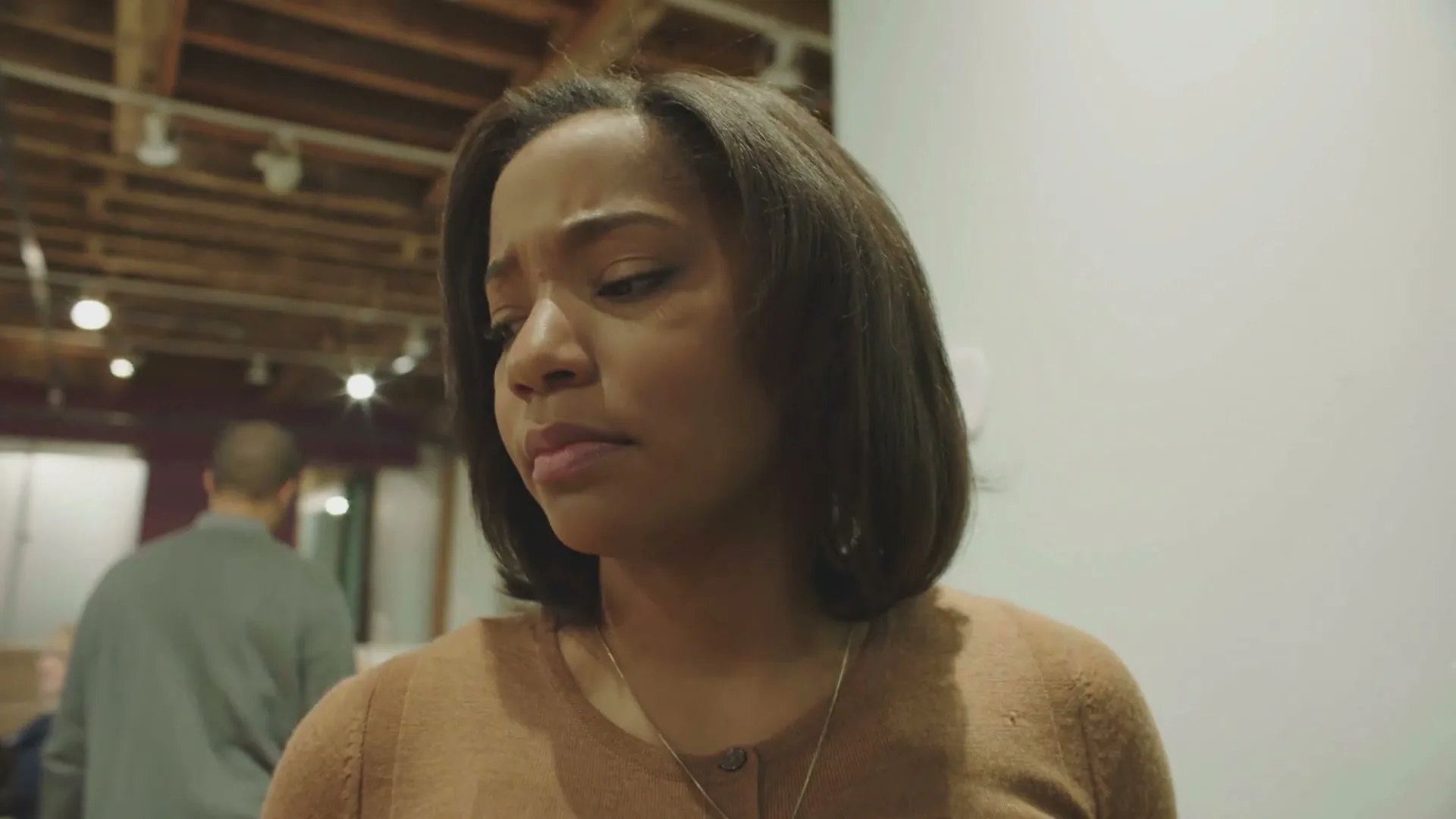 A woman with dark hair and a brown top stands in an office, looking down with a pained expression, as people walk past her in the background.