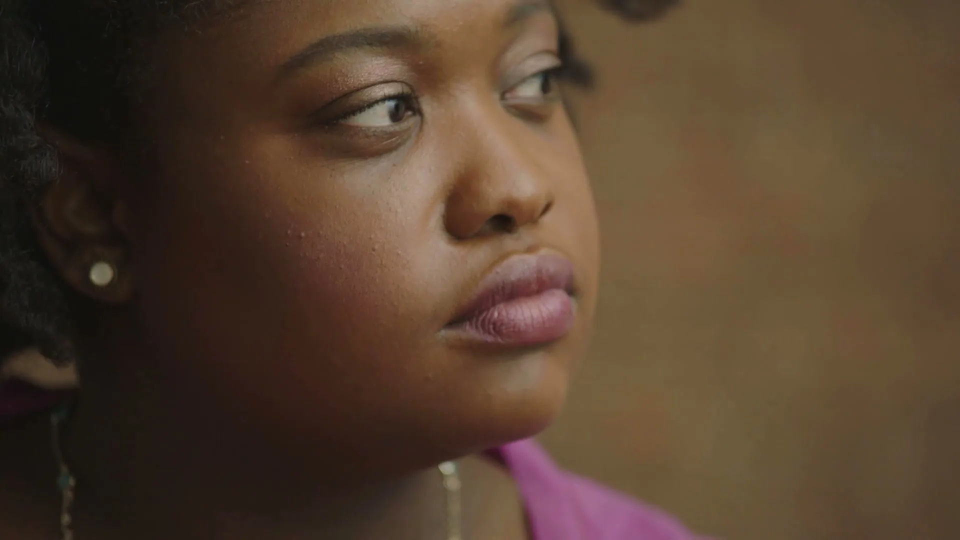 A young Black woman with dark hair and a magenta top looks off-camera with a neutral expression in a softly lit interior.