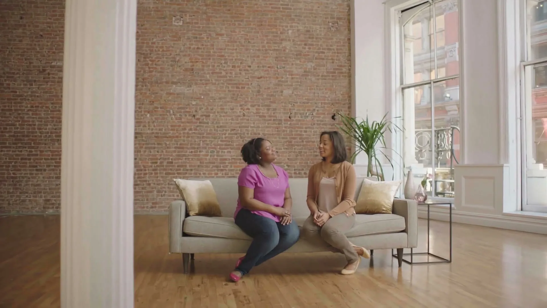 Two women sit on a couch in a spacious, sunlit loft with exposed brick walls and large windows, engaged in conversation.