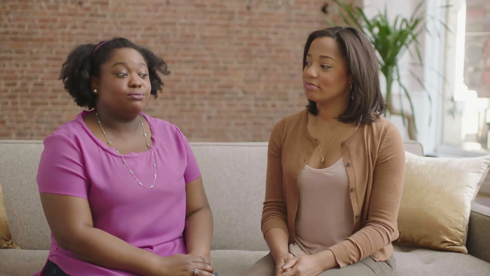 Two women sit on a couch, one speaking animatedly while the other listens intently in a brightly lit living room.