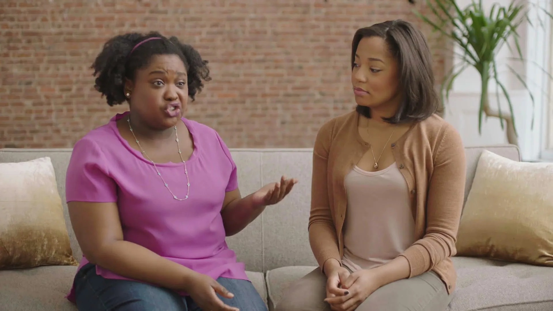 Two women sit on a couch, one speaking animatedly while the other listens intently in a living room.