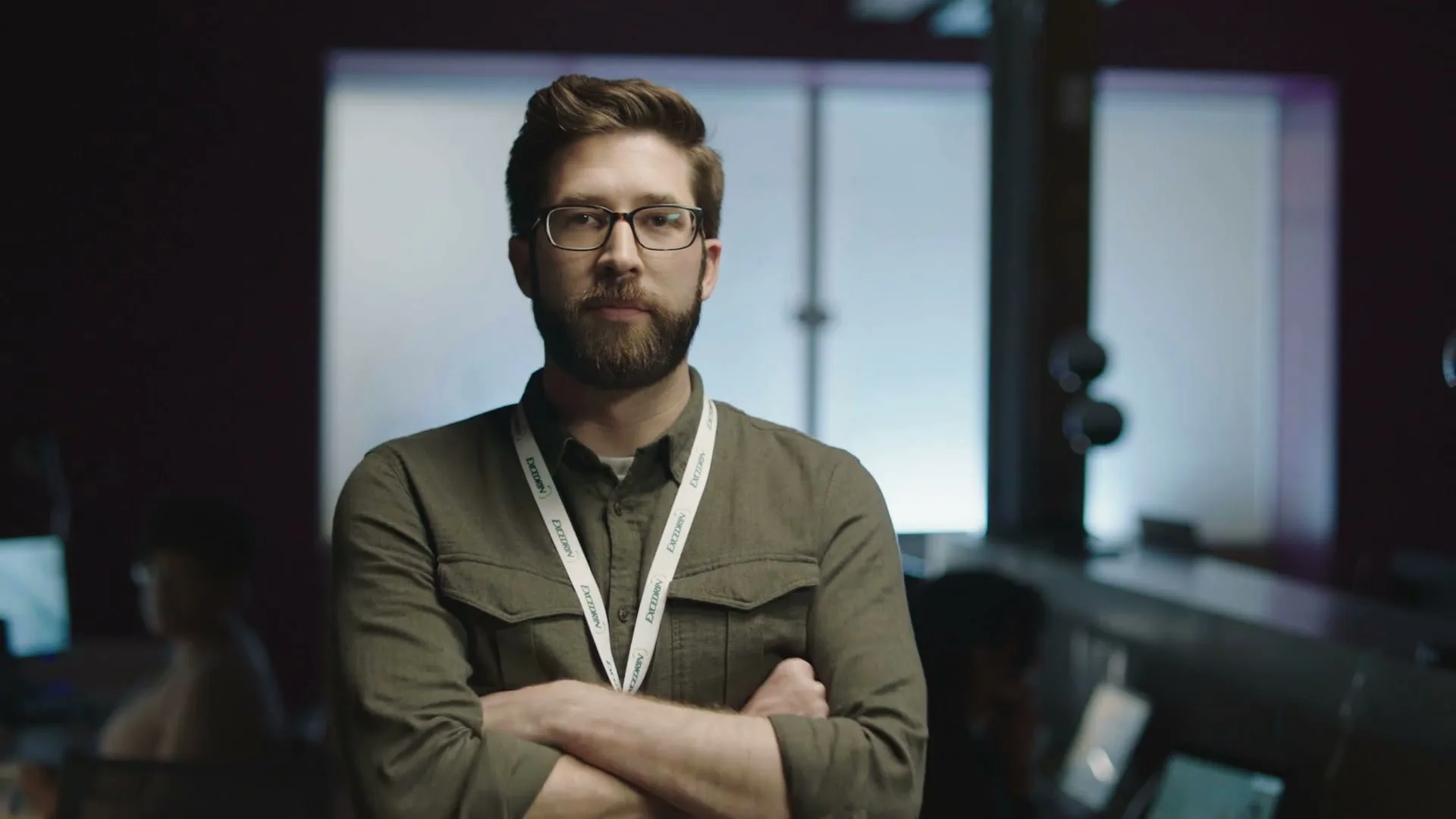 A man with a beard and glasses, wearing a green shirt and a lanyard, opens his eyes and crosses his arms in a dark office.